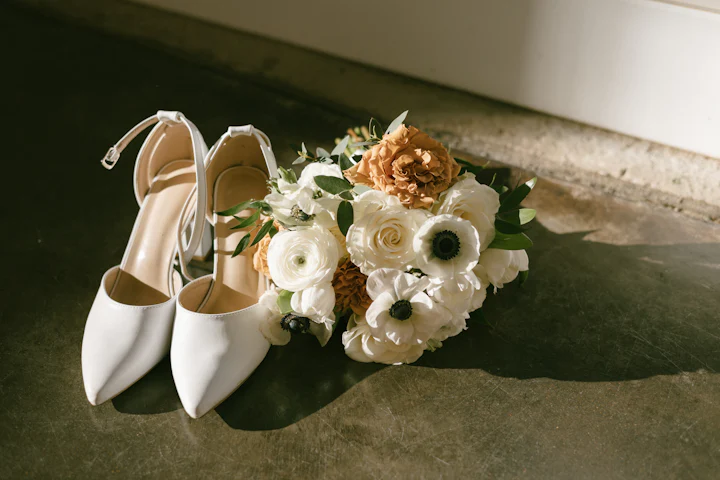 A bouquet of white anemones and cream roses resting beside a pair of white shoes.