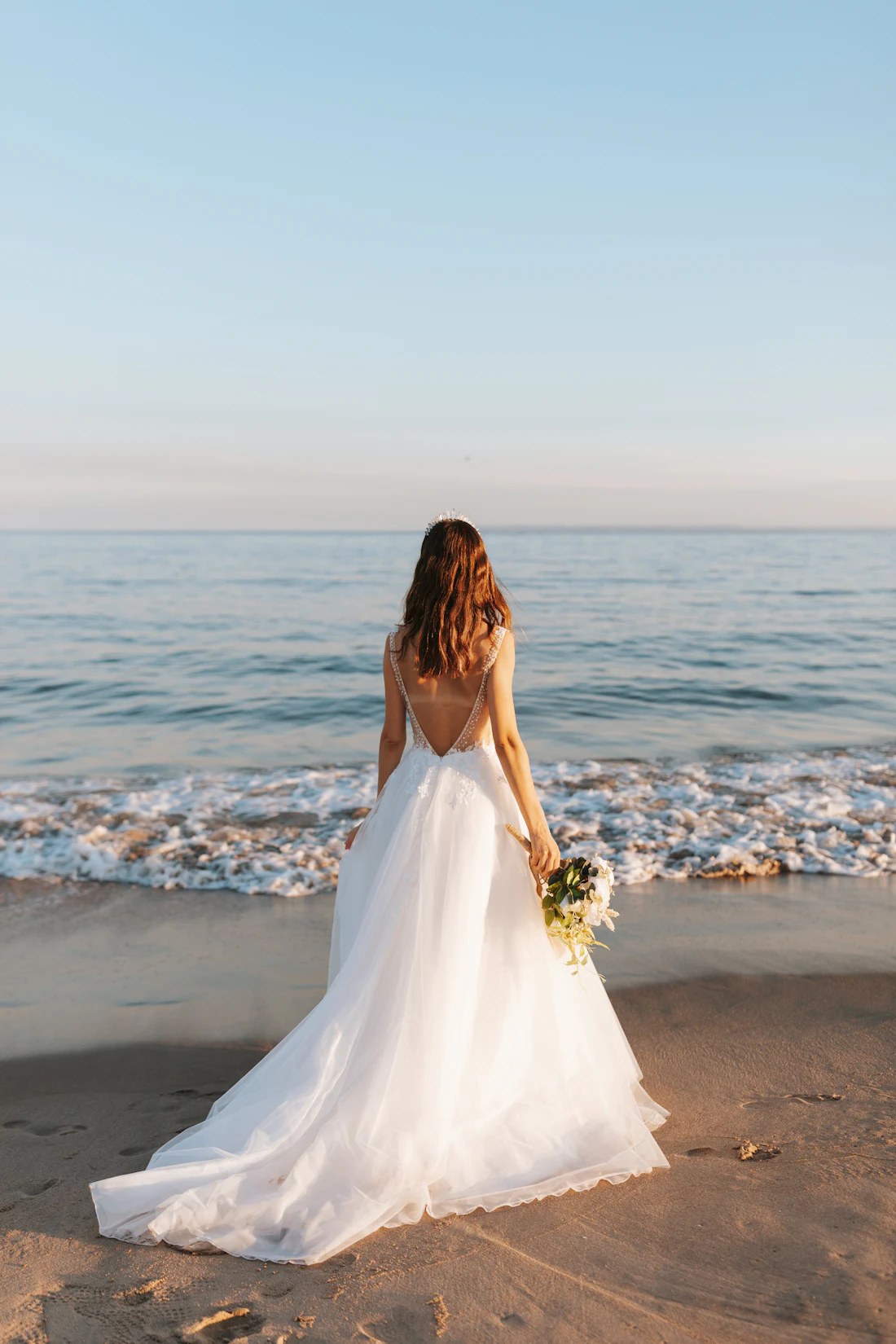 A bride in a low-backed gown standing on wet sand, facing a calm sea at low sun.