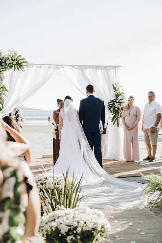 A couple exchanging vows at a white draped arch at the water&rsquo;s edge.