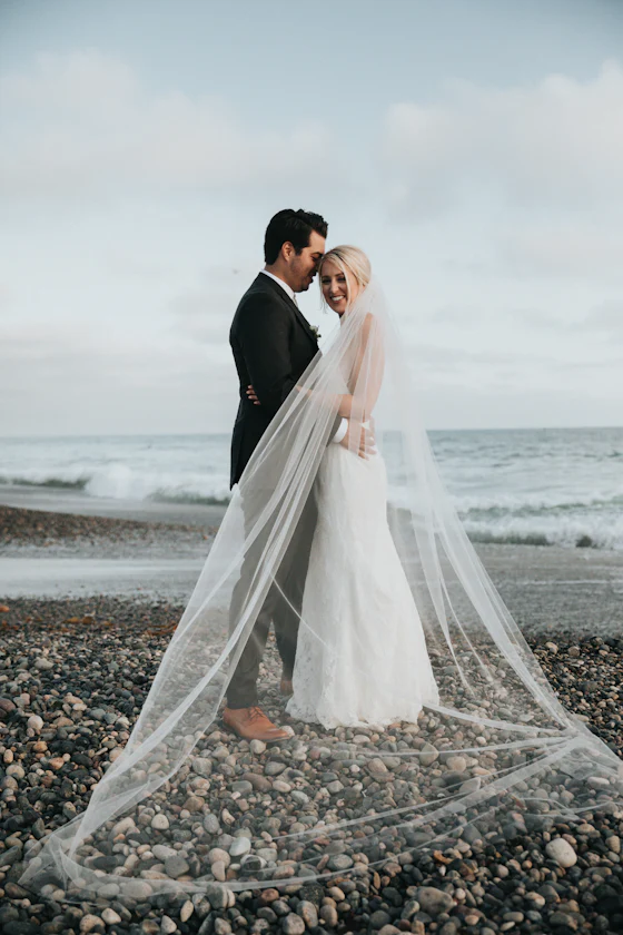 A couple standing on a pebble shore with a cathedral-length veil trailing behind them.