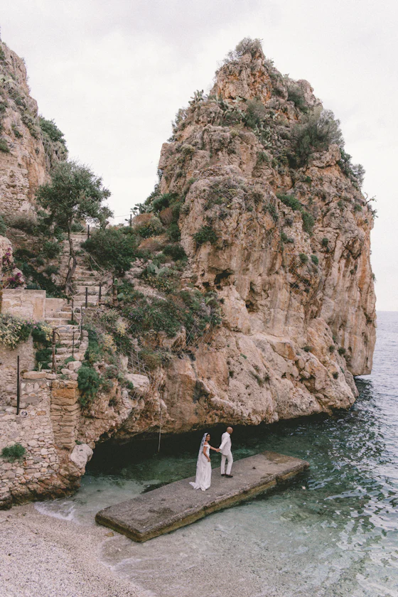 A couple standing on a small wooden dock beneath a rocky cliff.