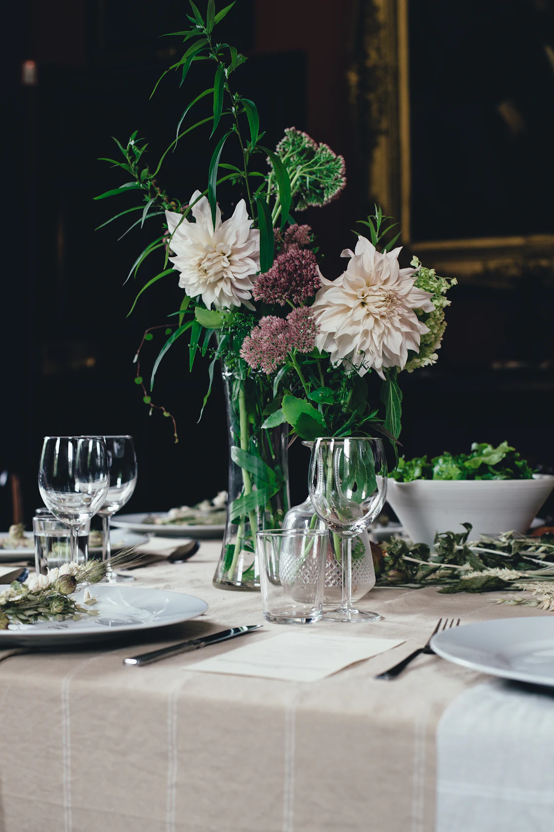 White and rust dahlias in low vases on a white-linen table with dark plates.