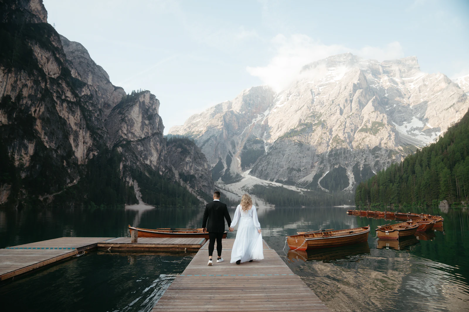 A bride and groom walking away along a wooden dock toward a mountain lake and jagged peaks.
