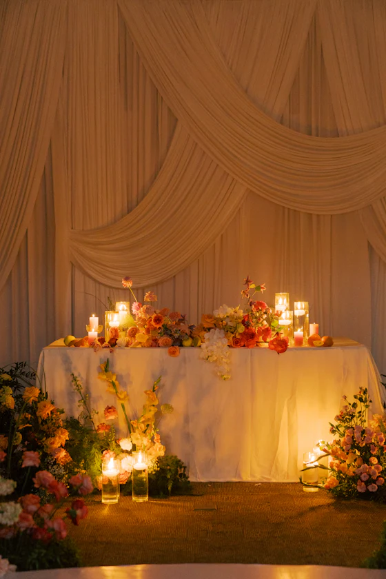 A sweetheart table against gathered silk drapery, banked with flowers and candles.