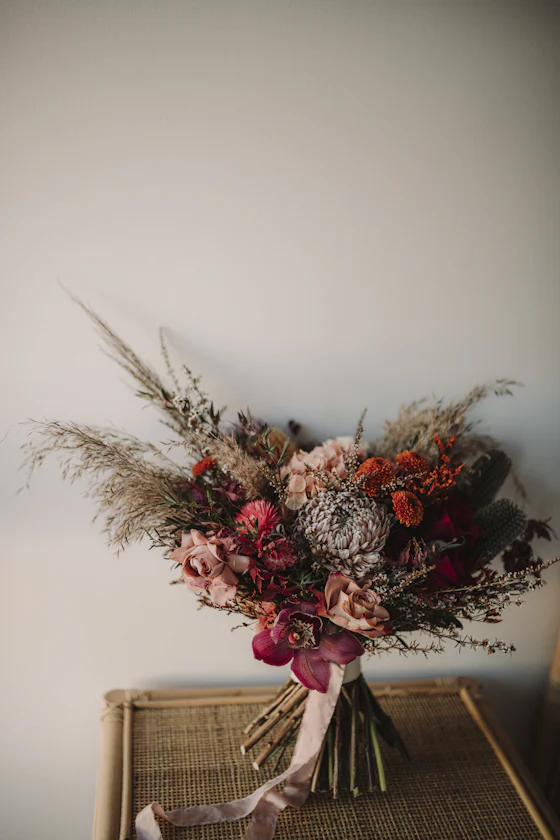 A loose bouquet of dried grasses and deep pink roses resting on a wicker chair.