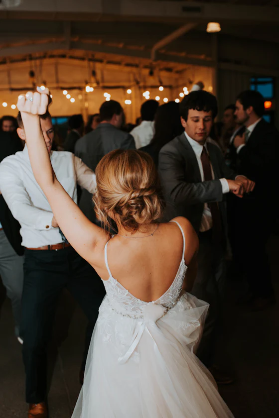 A bride in a low-backed gown dancing among guests beneath warm string lights.