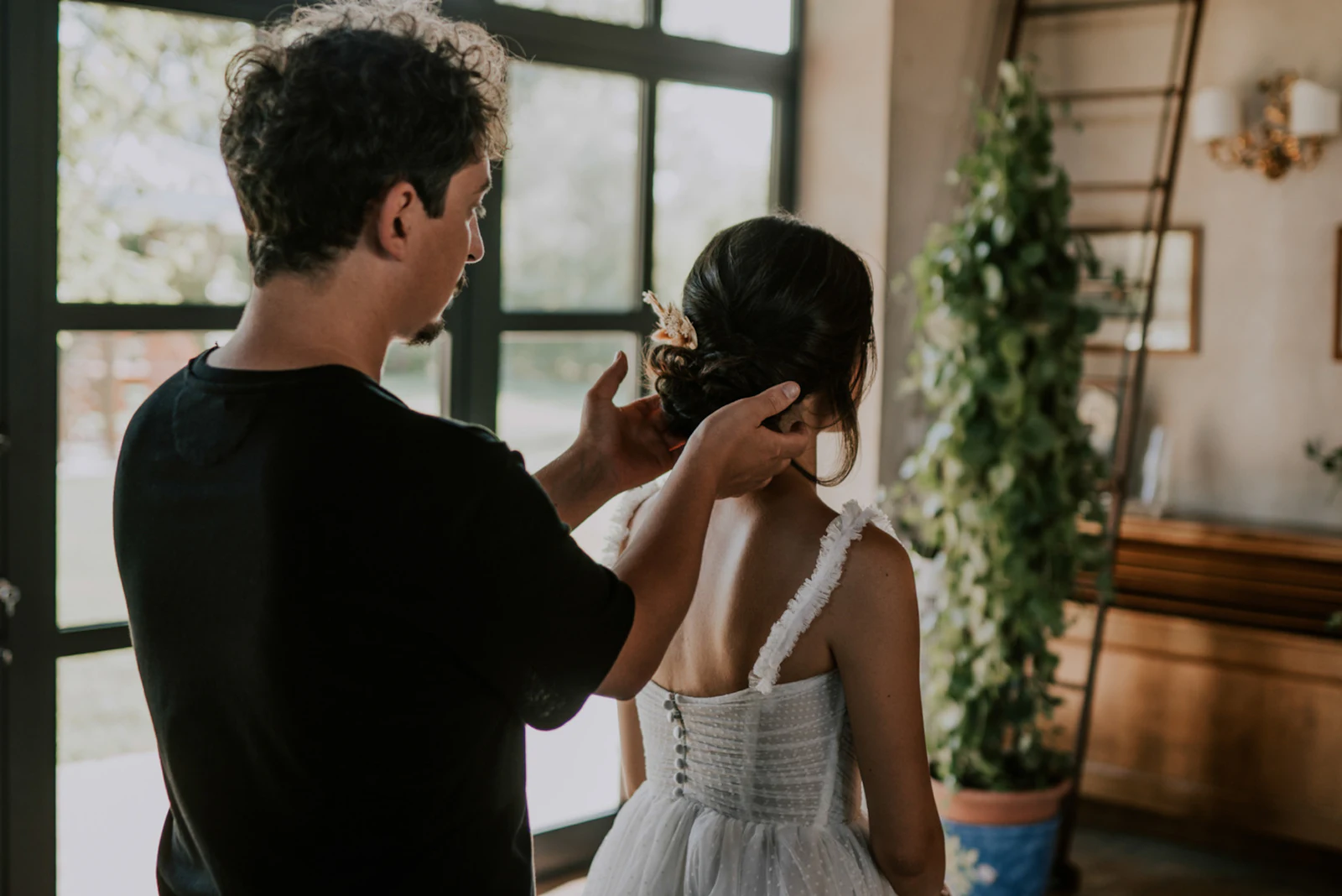 A groomsman helping a bride with the final buttons of her gown beside a tall window.