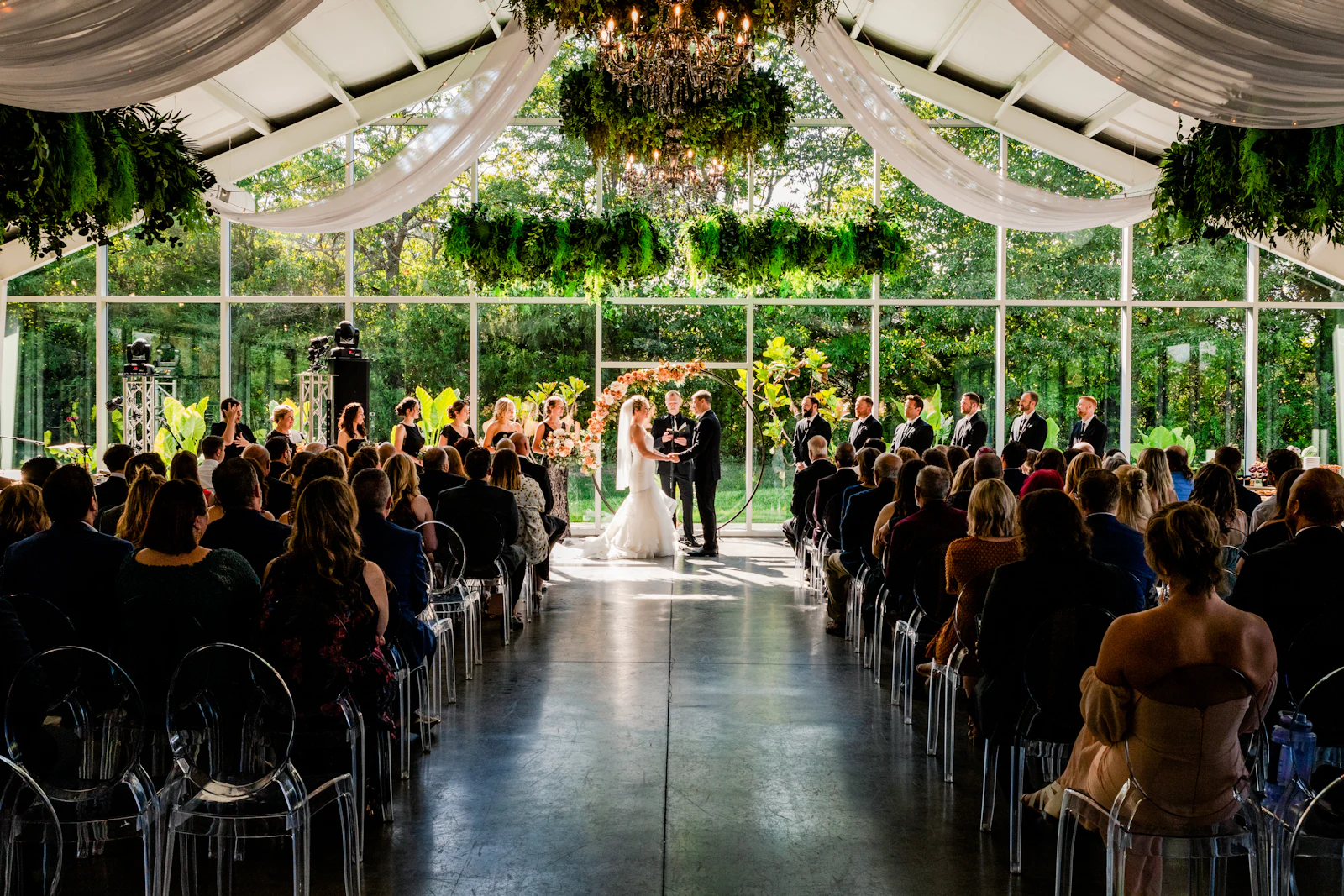 A couple at the end of a glass-roofed aisle beneath hanging greenery, guests seated on both sides.