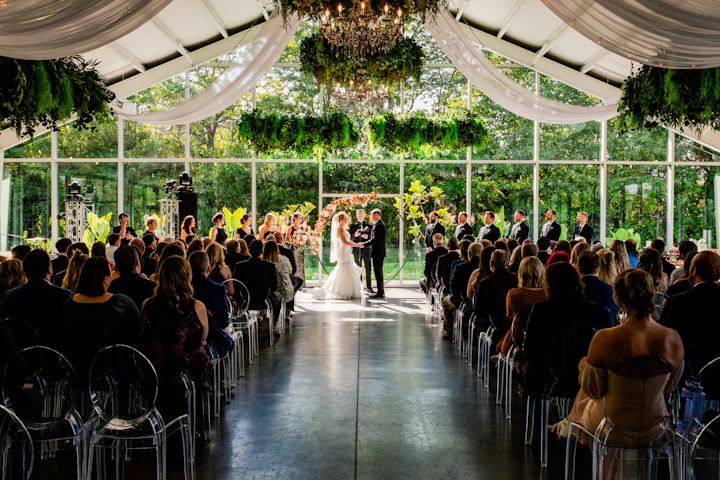 A couple at the end of a glass-roofed aisle beneath hanging greenery, guests seated on both sides.