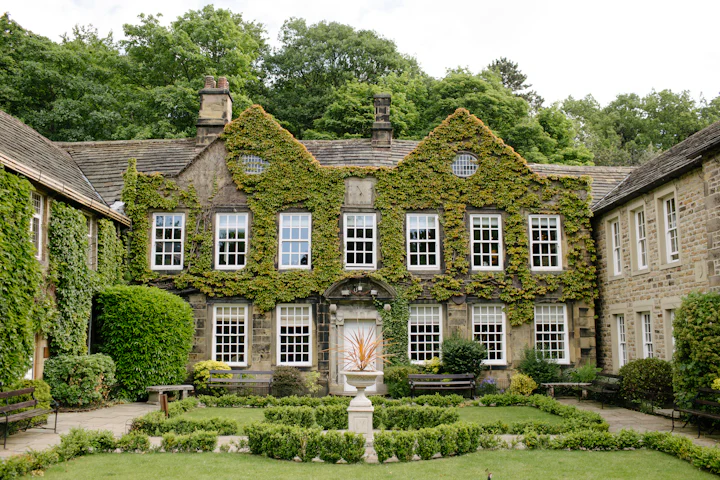 An ivy-covered manor house with tall white-framed windows, seen from its formal garden.