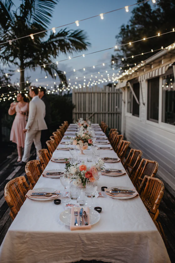 A long dinner table set under string lights and palms as dusk falls.