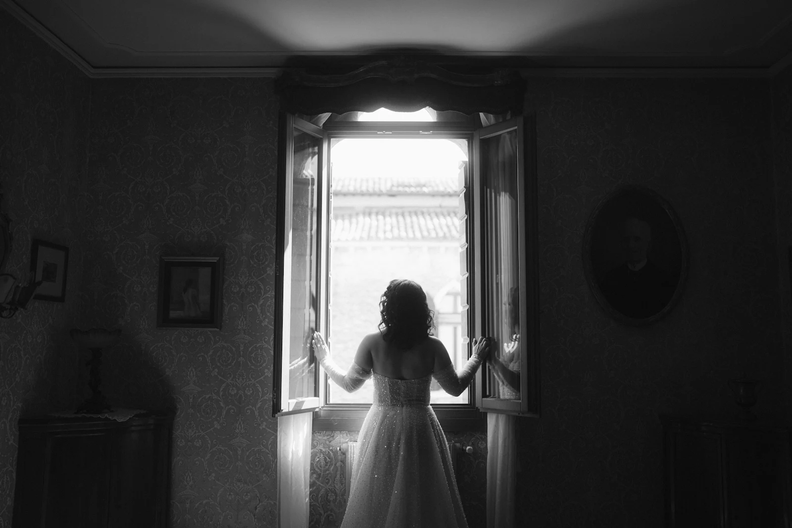 A bride in monochrome, standing at tall open windows looking out.